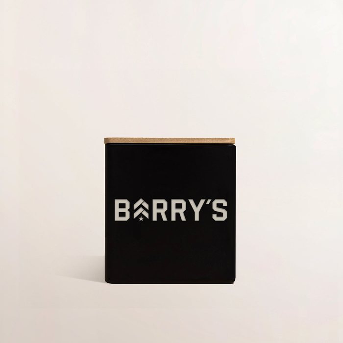 A black TAJA logo candle engraved for Barry's, on a pale studio backdrop