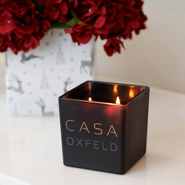 A square black candle tin with the brand name 'CASA OXFELD' in gold lettering, lit with two flames, sitting on a white marble countertop next to a white vase of red flowers.