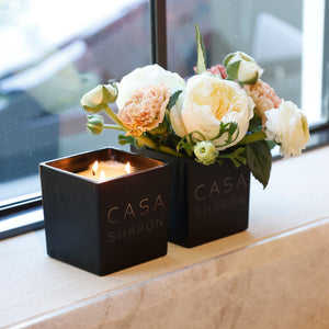 Two black square candles by CASA SHARON sit on a beige stone windowsill. One candle is lit and the other holds a bouquet of cream and peach roses with green leaves.