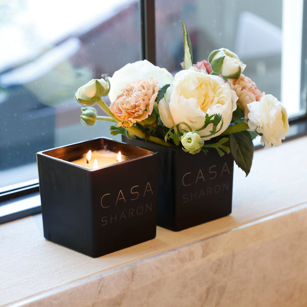 Two black square candles by CASA SHARON sit on a beige stone windowsill. One candle is lit and the other holds a bouquet of cream and peach roses with green leaves.