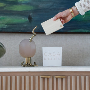 A hand wearing a white sleeve and gold bracelets holds a cream-colored rectangular card in front of a marble counter. On the counter are a frosted pink glass sculpture with a gold snake-like finial, a white square card reading 'CASA RHODES', and a beige ribbed cabinet with gold handles.