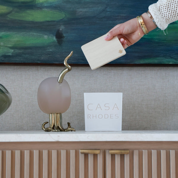 A hand wearing a white sleeve and gold bracelets holds a cream-colored rectangular card in front of a marble counter. On the counter are a frosted pink glass sculpture with a gold snake-like finial, a white square card reading 'CASA RHODES', and a beige ribbed cabinet with gold handles.
