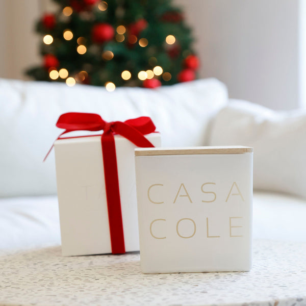 A white square Casa Cole candle tin with gold lettering stands on a speckled table next to a white gift box tied with a red ribbon, with a blurred Christmas tree in the background.