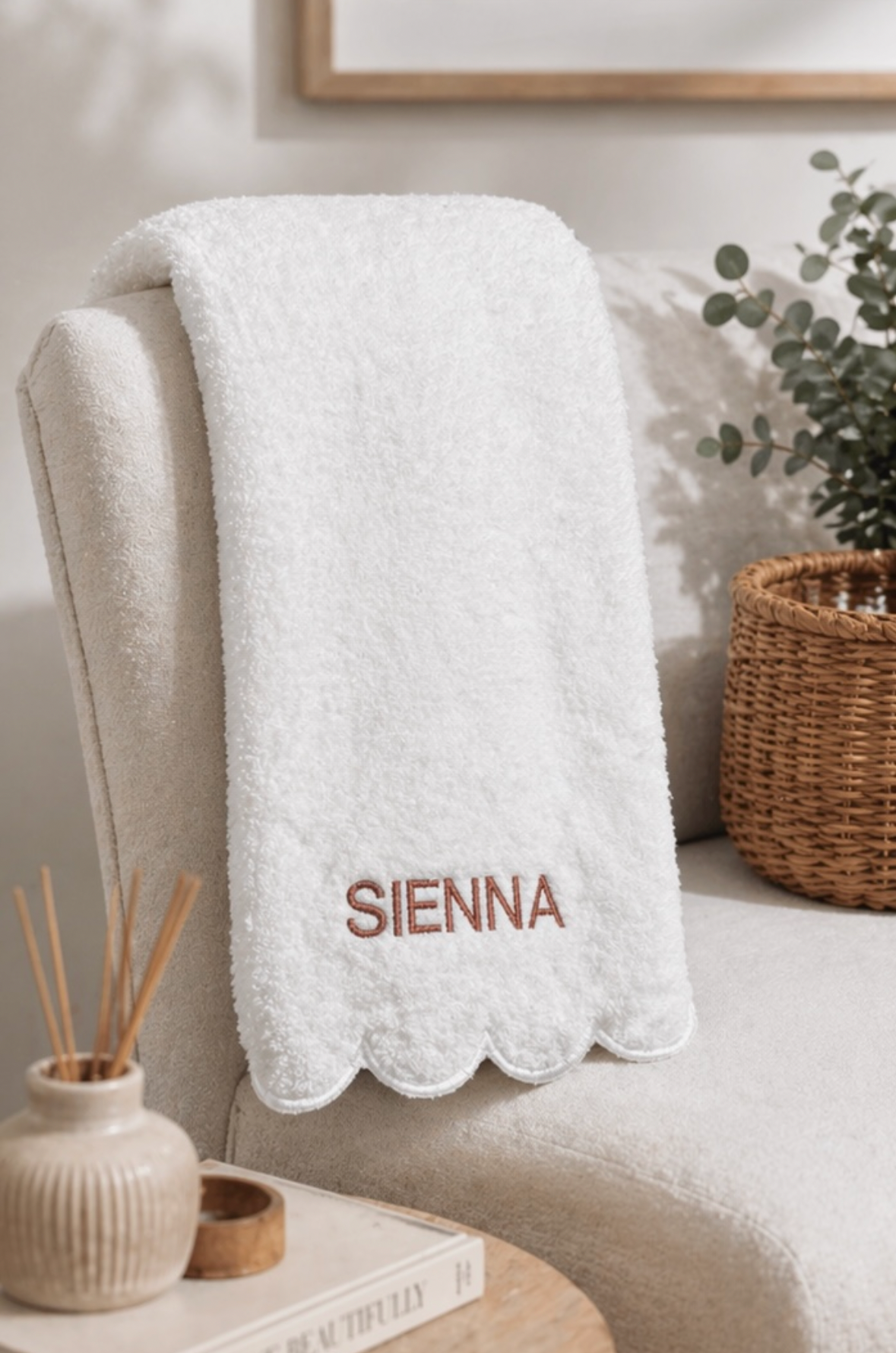 A white plush throw blanket with a scalloped edge and the name 'SIENNA' embroidered in brown on the front, draped over a cream-colored armchair in a styled room setting.