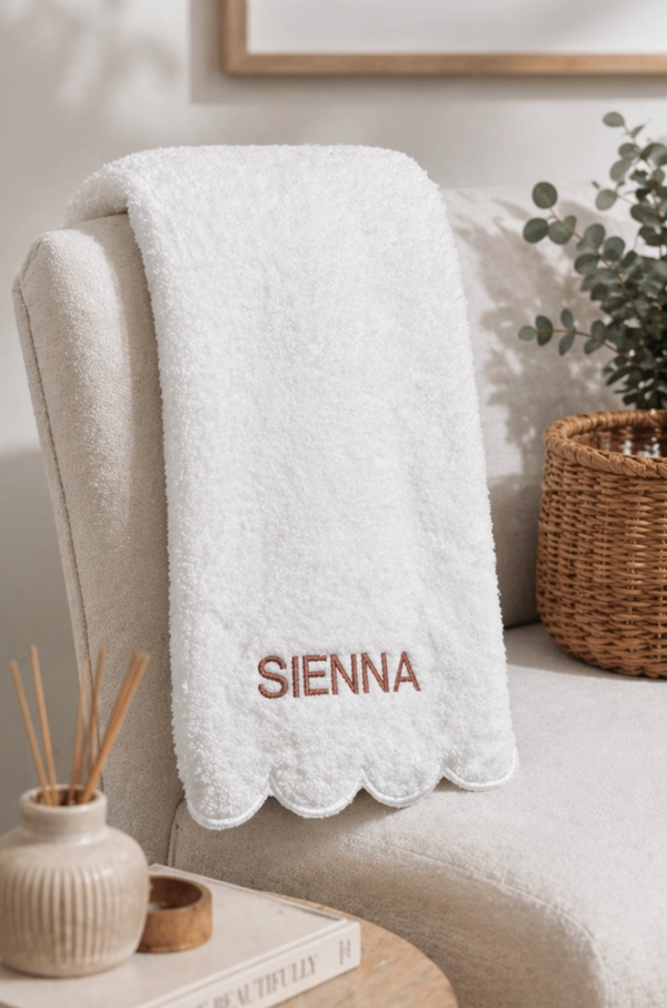 A white plush throw blanket with a scalloped edge and the name 'SIENNA' embroidered in brown on the front, draped over a cream-colored armchair in a styled room setting.