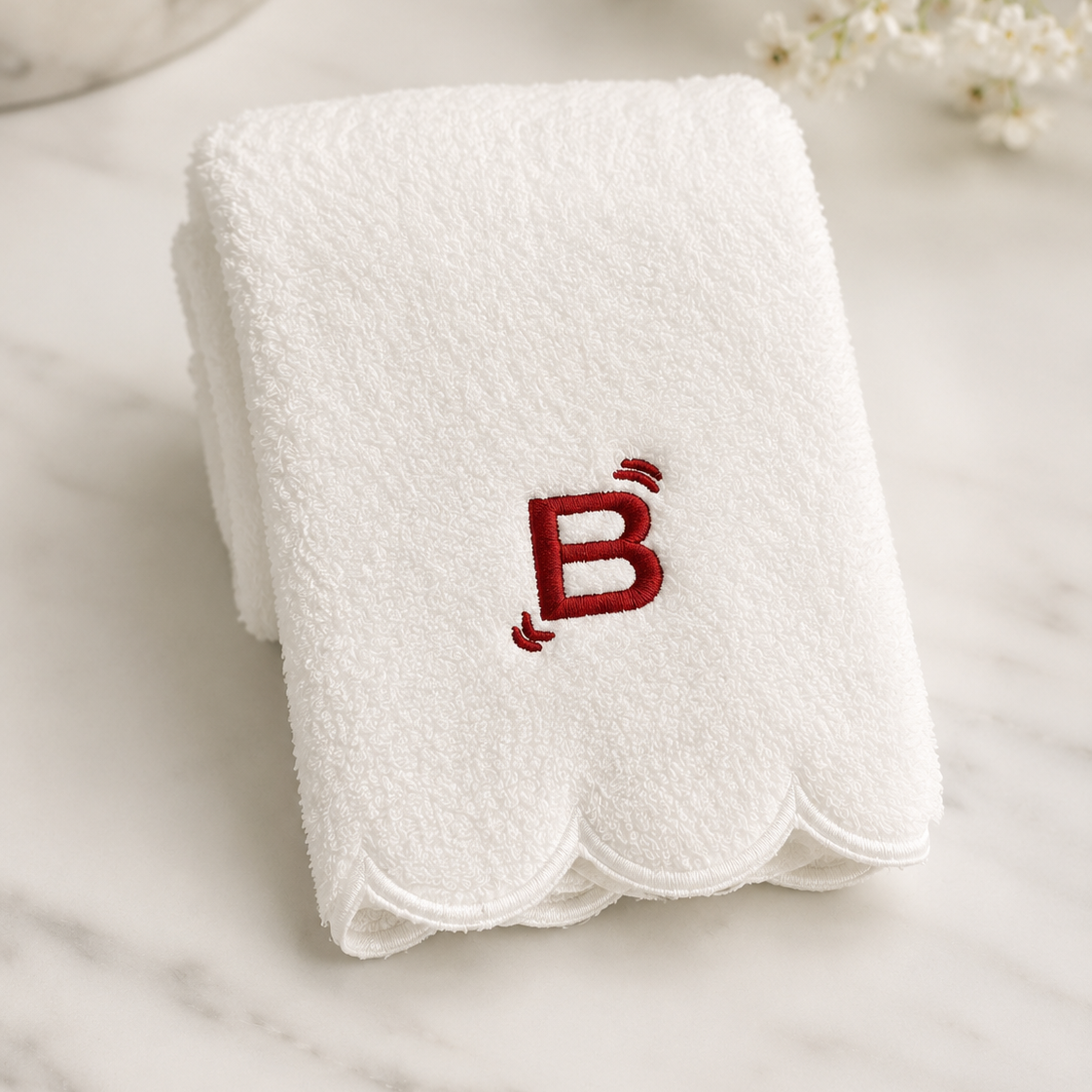 A folded white terry cloth hand towel with a scalloped edge and an embroidered red letter 'B' on the front, displayed on a light marble surface with small white flowers in the background.