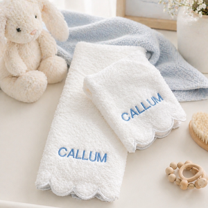A set of two white personalized baby towels with scalloped edges and blue embroidered 'CALLUM' text, styled on a white dresser next to a plush bunny toy, a light blue blanket, and baby accessories.