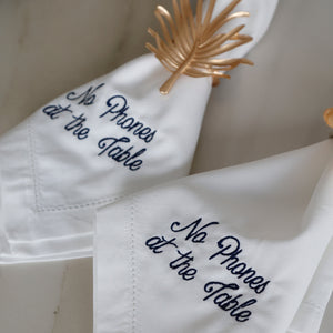 Two white napkins with the phrase 'No Phones at the Table' embroidered in navy blue script, styled on a light marble surface with a gold palm leaf napkin ring.