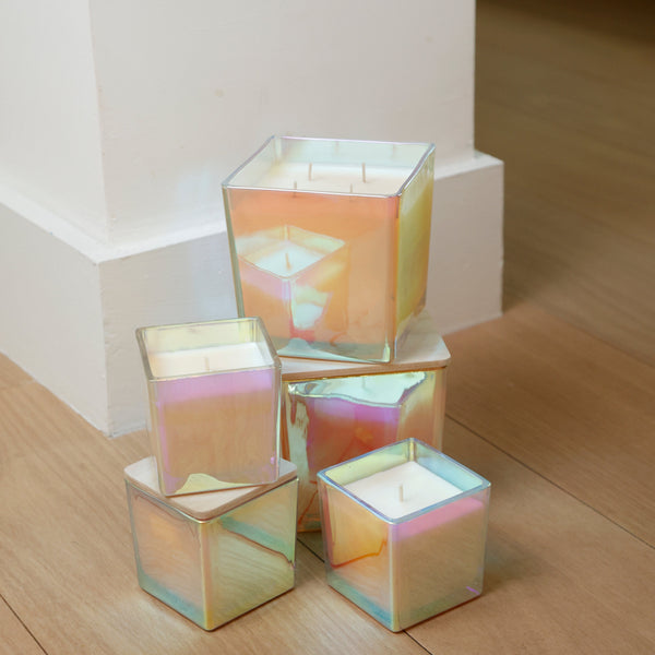 Four square iridescent glass candle holders with white candles inside, arranged on a light wood floor next to a white baseboard.
