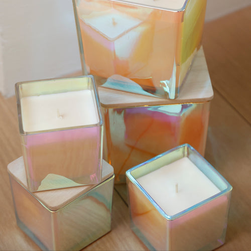 Four square glass candles with iridescent, holographic finishes in pastel pink, orange, yellow, and blue, arranged on a light wood floor against a white wall.