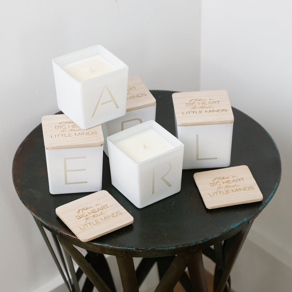 Five white square decorative boxes with light wood lids arranged on a dark round table. Each lid is engraved with the phrase 'BIG HEART LITTLE MINDS' and one box shows a large gold letter, with the others showing partial letters.