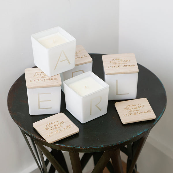 Three white square candle jars with light wood lids stacked on a dark round table, alongside matching wooden coasters. The lids are engraved with the name 'EARL' and a quote from 'The Little Things'.