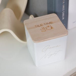 A white square ceramic candle tin with a light natural wood lid engraved with 'TILE TIME' and two hexagons, featuring 'Game Night' in gold script on the front, styled on a white surface next to books and a beige sculptural object.