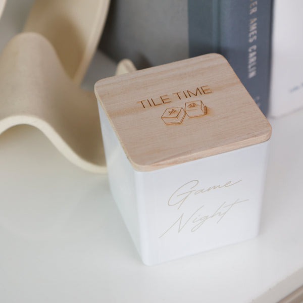 A white square ceramic candle tin with a light natural wood lid engraved with 'TILE TIME' and two hexagons, featuring 'Game Night' in gold script on the front, styled on a white surface next to books and a beige sculptural object.