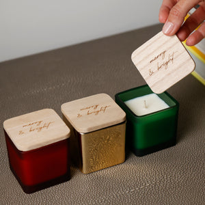 Three square glass candles in red, gold, and green with light wood lids engraved with 'many & bright', arranged on a textured surface with a hand lifting one lid.
