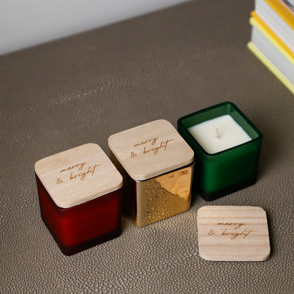Three square glass candles in red, gold, and green with light wood lids engraved with 'many & bright', arranged on a textured surface with a hand lifting one lid.