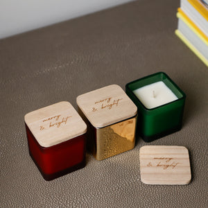 Three square glass candle jars with wooden lids are arranged on a textured brown surface. One jar is red, one is metallic gold, and one is dark green with white wax visible.