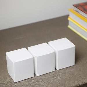Three plain white cube-shaped boxes arranged in a row on a textured brown surface, with a blurred stack of books in the background.