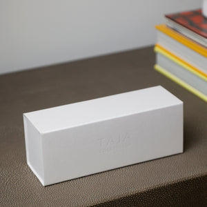 A minimalist white rectangular product box with embossed 'TAJA' branding, resting on a textured brown leather surface with a blurred stack of colorful books in the background.