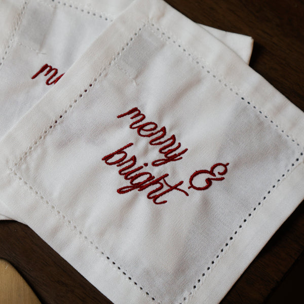 A close-up of white embroidered napkins with red script reading 'merry & bright' on a dark wooden surface.
