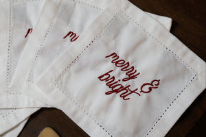 A close-up of white embroidered napkins with red script reading 'merry & bright' on a dark wooden surface.