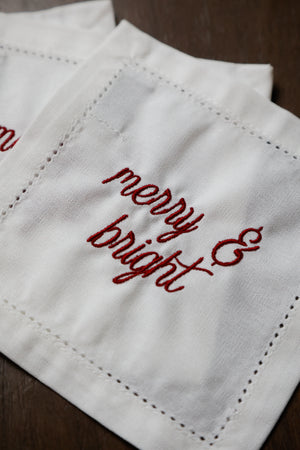 A close-up of a white embroidered cloth napkin with the words 'merry & bright' stitched in red cursive, resting on a dark wooden surface.