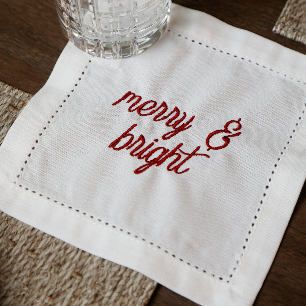 A close-up of white embroidered napkins with red script reading 'merry & bright' on a dark wooden surface.