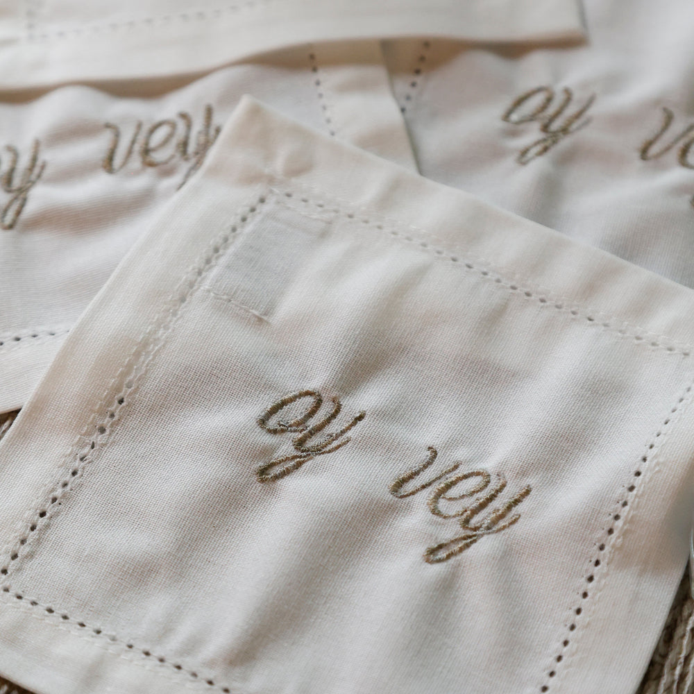 Close-up of several white embroidered napkins with cursive script reading 'oy vey' and stitched border details, arranged on a textured beige surface.