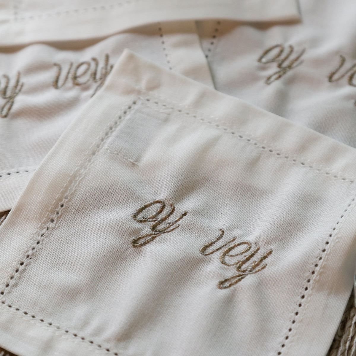 Close-up of several white embroidered napkins with cursive script reading 'oy vey' and stitched border details, arranged on a textured beige surface.