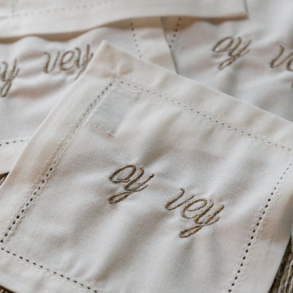 Close-up of several white embroidered napkins with cursive script reading 'oy vey' and stitched border details, arranged on a textured beige surface.