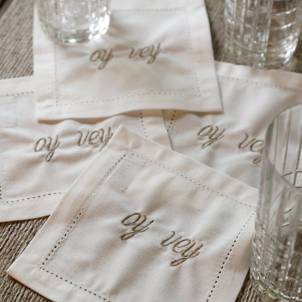 A set of square off-white linen napkins with embroidered cursive text reading 'of rey' and stitched border details, arranged on a woven placemat with clear drinking glasses around them.
