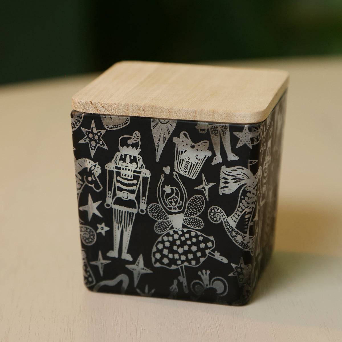 A square wooden box with a light natural wood lid and a black side panel decorated with white whimsical illustrations of nutcrackers, fairies, stars, and cups, shown on a light wood surface.