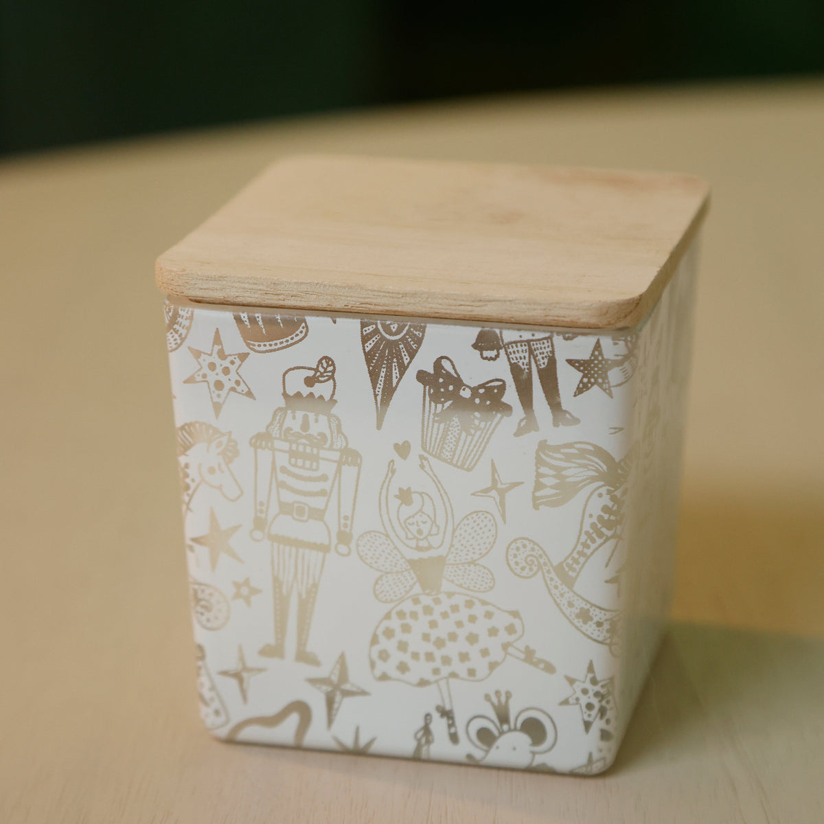A square ceramic container with a light wood lid, decorated with a white body and brown whimsical illustrations of nutcrackers, stars, and patterns, sitting on a light wooden surface.