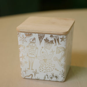 A square ceramic container with a light wood lid, decorated with a white body and brown whimsical illustrations of nutcrackers, stars, and patterns, sitting on a light wooden surface.