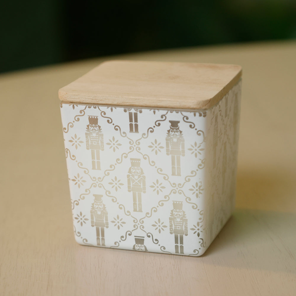 A square ceramic container with a light natural wood lid, decorated with a white and beige nutcracker pattern, sitting on a light wood surface with a dark green blurred background.