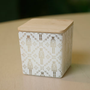 A square ceramic container with a light natural wood lid, decorated with a white and beige nutcracker pattern, sitting on a light wood surface with a dark green blurred background.