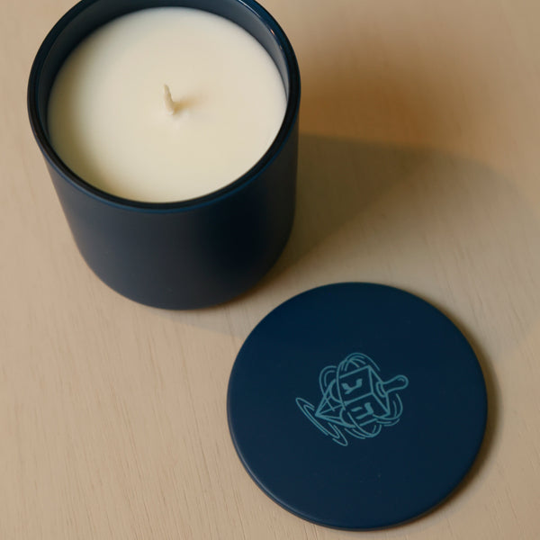 An open dark blue candle jar with white wax sits on a light wood surface, with the matching lid placed beside it. The lid features a light blue geometric line-art logo.