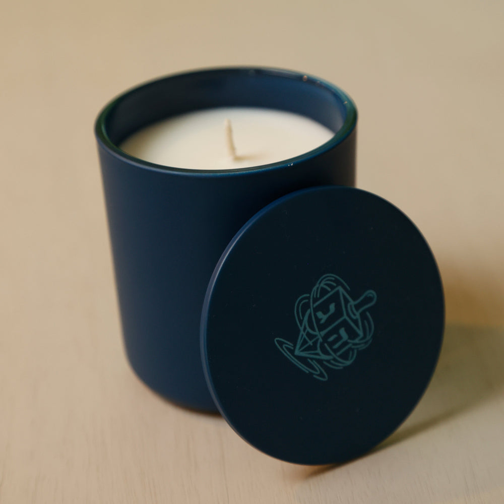 An open dark blue candle jar with white wax sits on a light wood surface, with the matching lid placed beside it. The lid features a light blue geometric line-art logo.