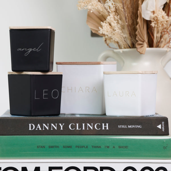 Four square metal candle tin boxes in black and white with gold lettering, stacked on top of books in a styled home decor setting with a ceramic vase of dried flowers in the background.