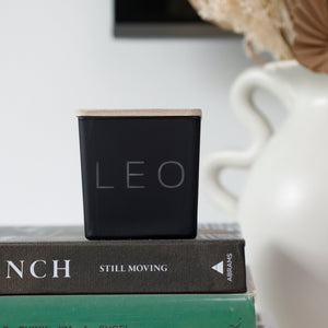 A square black candle with a light wood top and the word 'LEO' printed on the front, styled on top of two books next to a white ceramic vase with dried flowers.