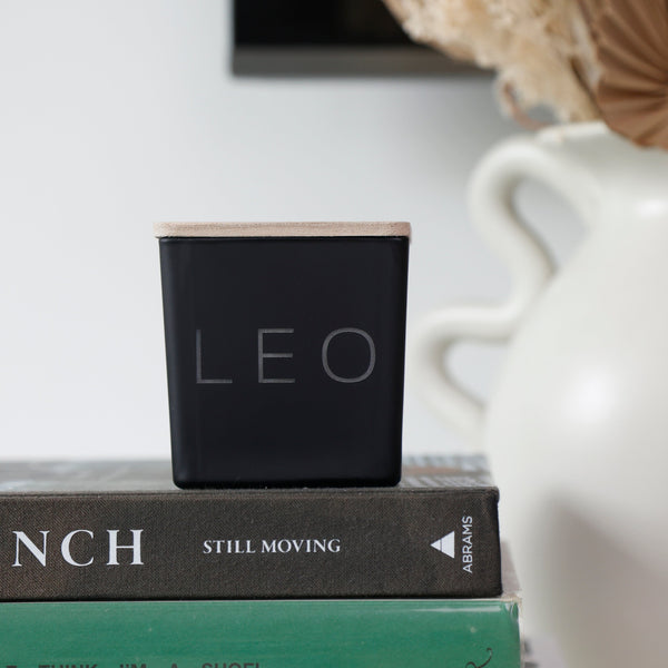 A square black candle with a light wood top and the word 'LEO' printed on the front, styled on top of two books next to a white ceramic vase with dried flowers.