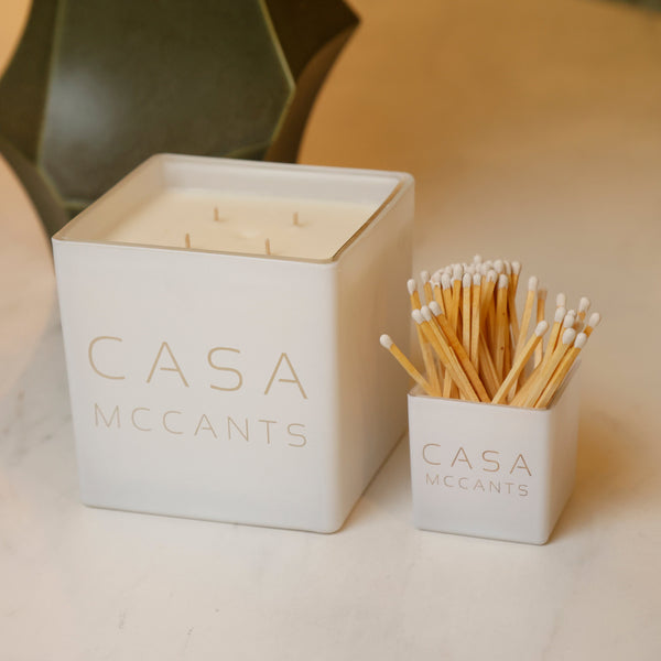 A Casa McCants soy candle in a white square container next to a small white holder filled with wooden matches, both branded with the Casa McCants name, set on a light marble surface.