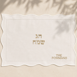 A square white fabric mat with a scalloped wavy border, featuring gold embroidered text in Hebrew and English reading 'THE FORMANS'  displayed flat on a beige textured surface with soft leaf shadows.