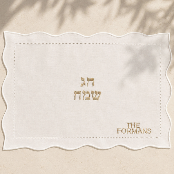 A square white fabric mat with a scalloped wavy border, featuring gold embroidered text in Hebrew and English reading 'THE FORMANS'  displayed flat on a beige textured surface with soft leaf shadows.