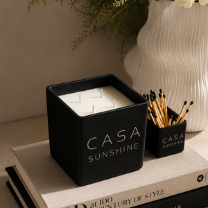 A black square Casa Sunshine candle jar with white wax sits on top of stacked books, next to a small black match holder with wooden matches. A white textured vase with flowers is in the background.