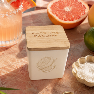A white square jar candle with a light wood lid engraved with 'PASS THE PALOMA' sits on a pink stone tray, surrounded by a pink cocktail, grapefruit, lime, and a small dish of salt in a styled setting.
