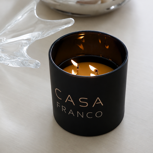 A black double-wick candle in an black glass jar with the brand name 'CASA FRANCO' engraved, set on a light beige surface with a blurred silver bowl in the background.