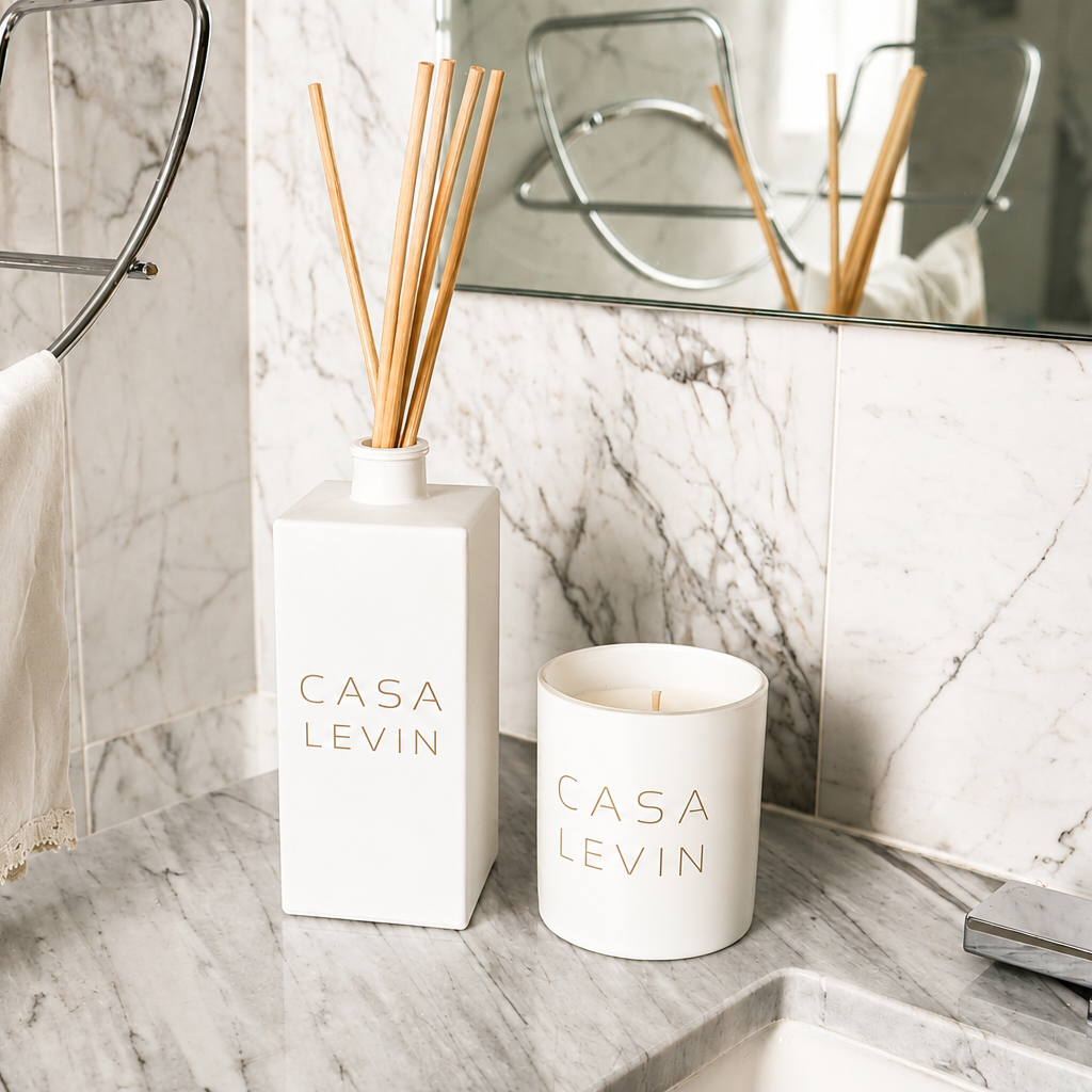 A Casa Levin reed diffuser and matching candle set on a marble bathroom counter, with a towel and mirror in the background.