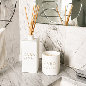 A Casa Levin reed diffuser and matching candle set on a marble bathroom counter, with a towel and mirror in the background.
