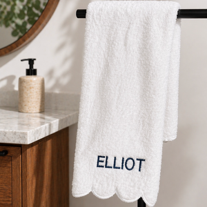 A white textured towel with scalloped edges and the name 'ELLIOT' embroidered in navy blue, hanging on a black rack in a bathroom setting.